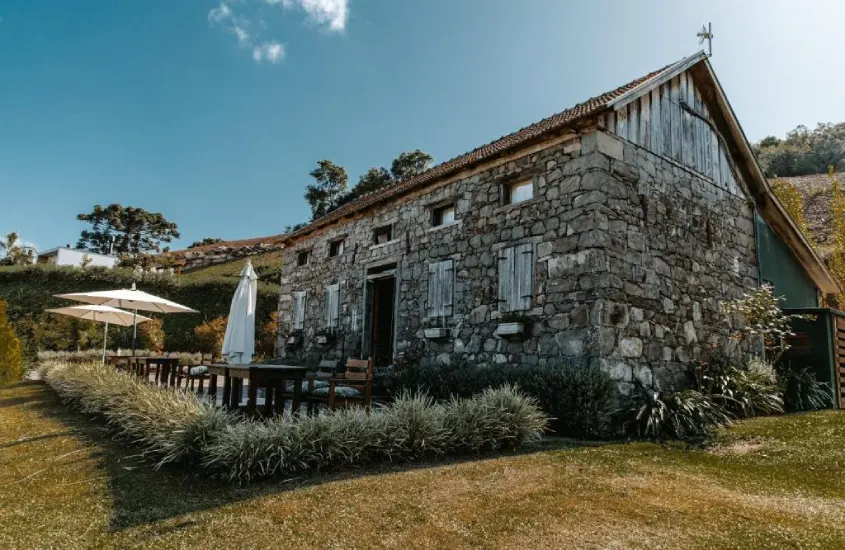 Fachada de um hotel vinícola com paredes de pedra, mesas, cadeiras, plantas ao redor e guarda-sóis