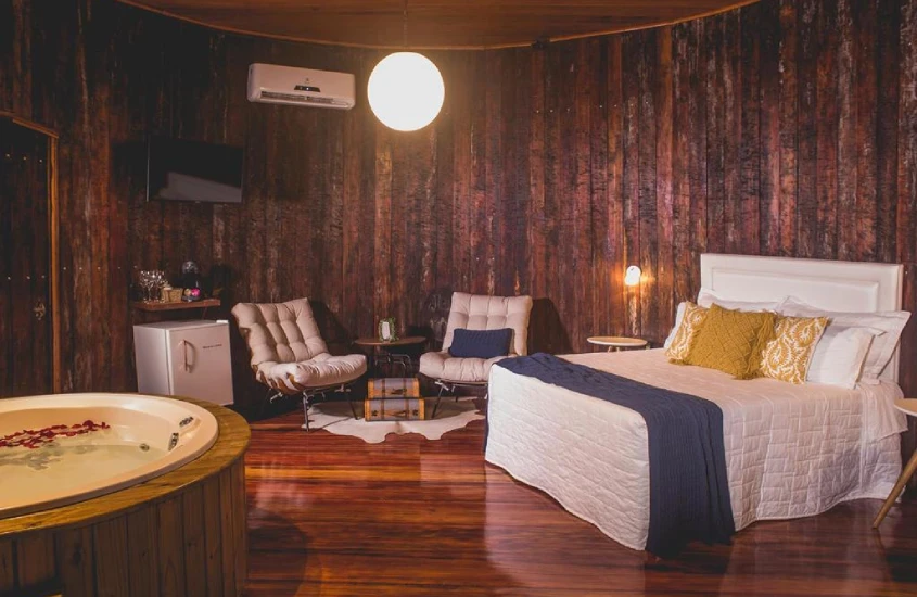 Interior de uma cabana com cama de casal, banheira de hidromassagem, cadeiras acolchoadas, TV e piso de madeira