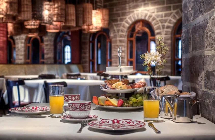 Mesa de café da manhã com frutas, suco, café, pães e mesas ao redor