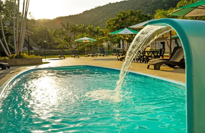 Em um final de tarde, área de lazer de hotel com piscina, espreguiçadeiras, guarda-sóis e árvores ao redor