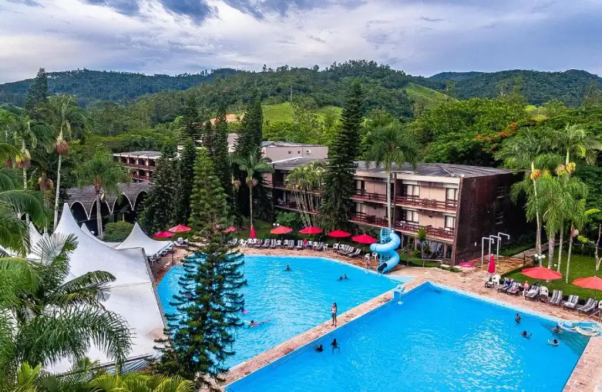 Em um dia nublado, área de lazer de hotel com piscinas, espreguiçadeiras, guarda-sóis, mesas, cadeiras, tendas e árvores ao redor