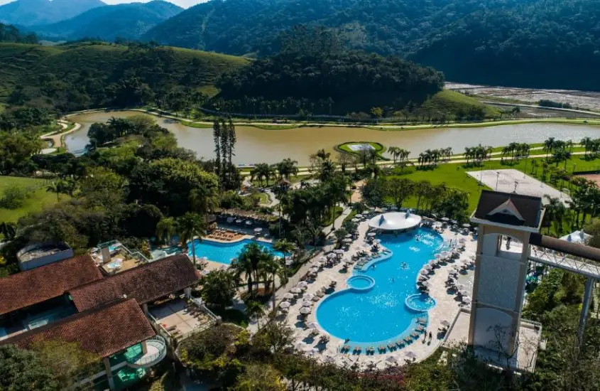 Visão aérea de hotel com lago do lado, piscinas, espreguiçadeiras, árvores e plantas ao redor