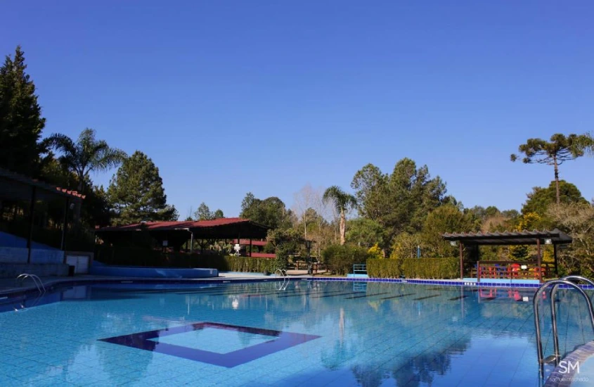 Em um dia de sol, área de lazer de hotel com piscina, espreguiçadeiras, gazebo de madeira e árvores ao redor