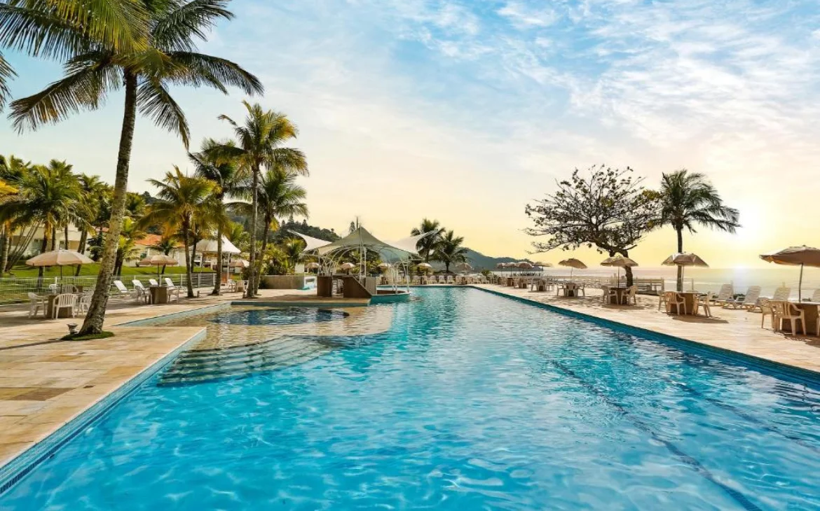 Em um dia de sol, área de lazer com piscina, espreguiçadeiras, praia do lado, árvores e plantas decorativas