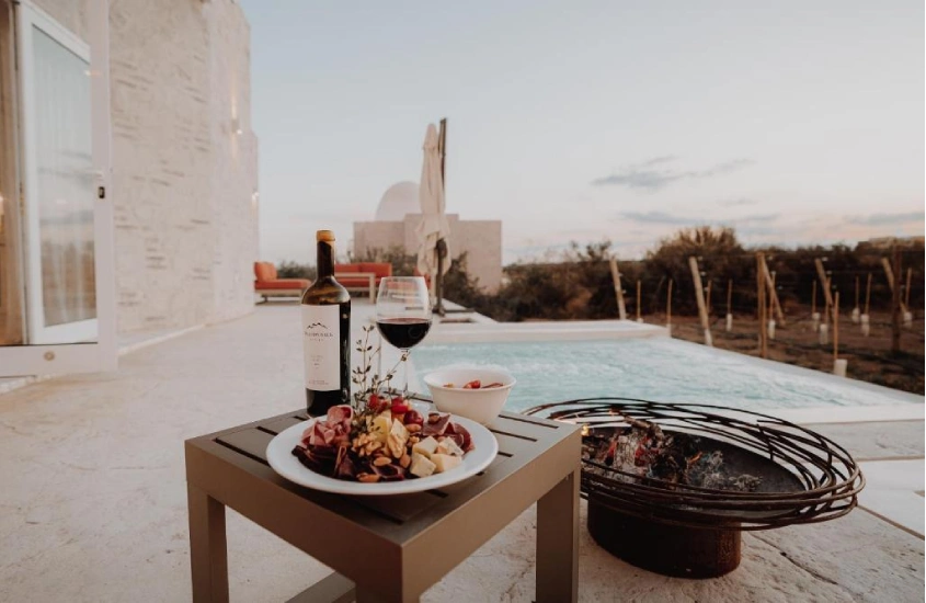 Em um final de tarde, área de lazer de um hotel com piscina fogueira, mesa de frios com vinho e árvores no fundo