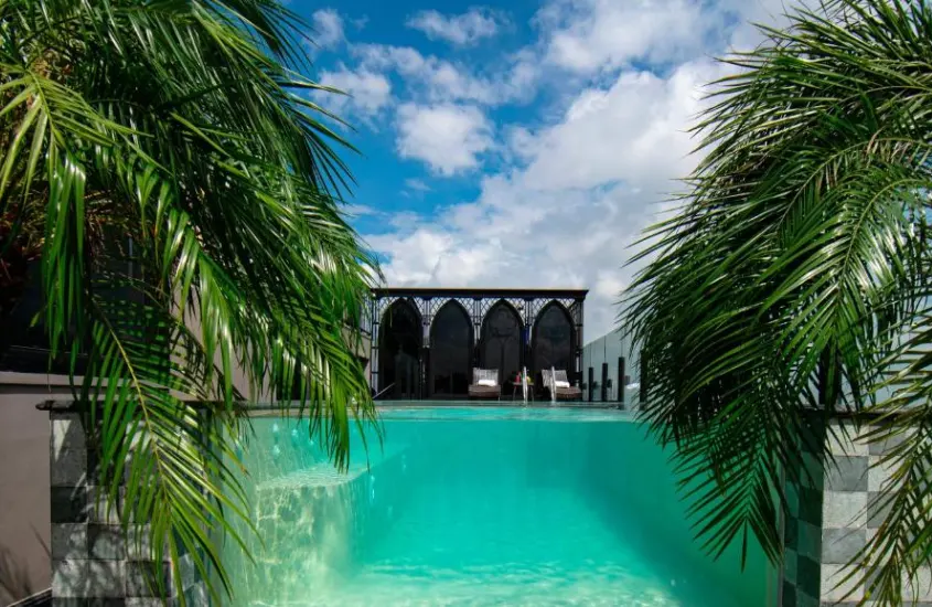 Em um dia de sol, área de lazer de hotel com piscina, tendas, espreguiçadeiras e árvores ao redor