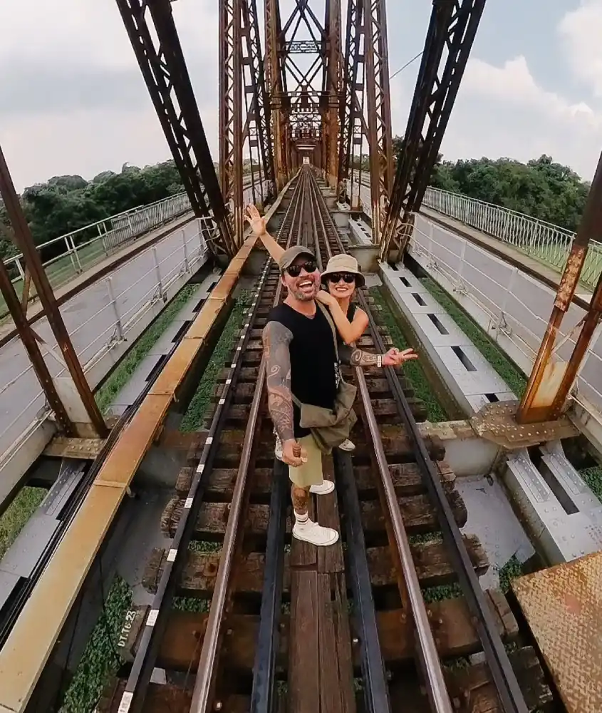 Vagner e Bárbara em trilho de trem com lago embaixo e árvores no caminho