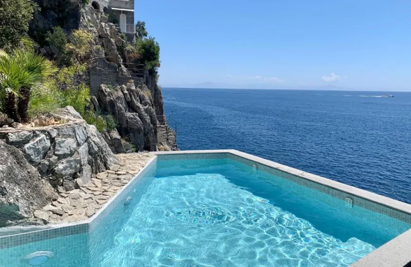 Em um dia de sol, área de lazer de hotel com piscina, paredões de pedra e mar do lado