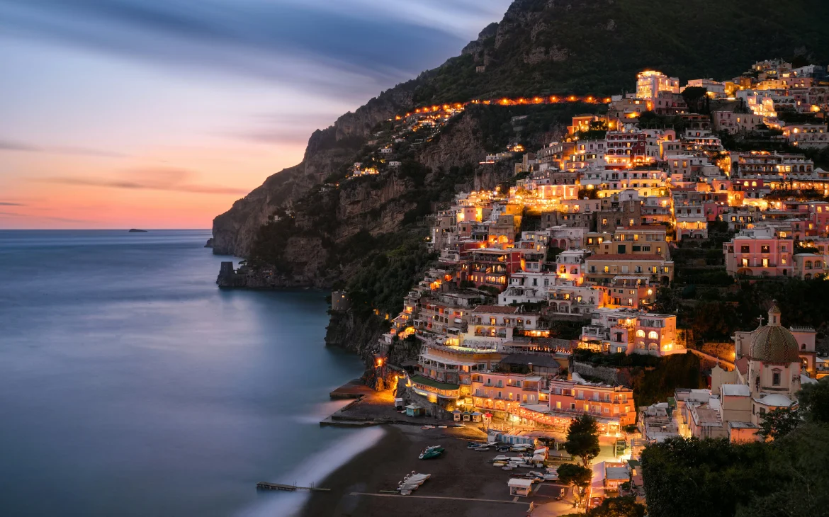 Durante o entardecer, paisagem da costa amalfitana com casinhas iluminadas e praia do lado com paredões de pedra