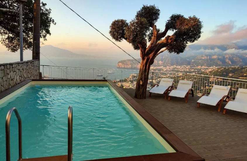 Durante o entardecer, piscina com espreguiçadeiras, árvores ao redor e vista da cidade