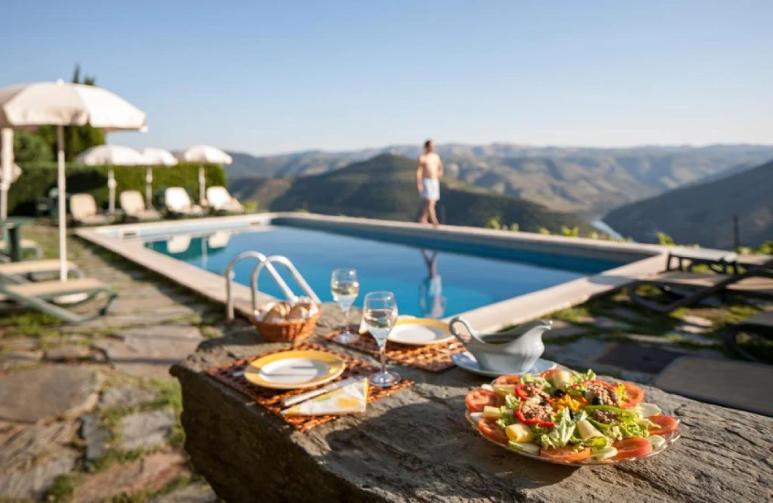 Em um dia de sol, área de lazer de hotel com piscina, espreguiçadeiras, mesa posta, guarda-sóis e montanhas ao redor