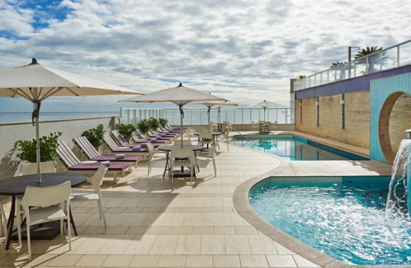 Em um dia de sol, área de lazer de hotel com piscinas, espreguiçadeiras, guarda-sóis e paisagem da praia