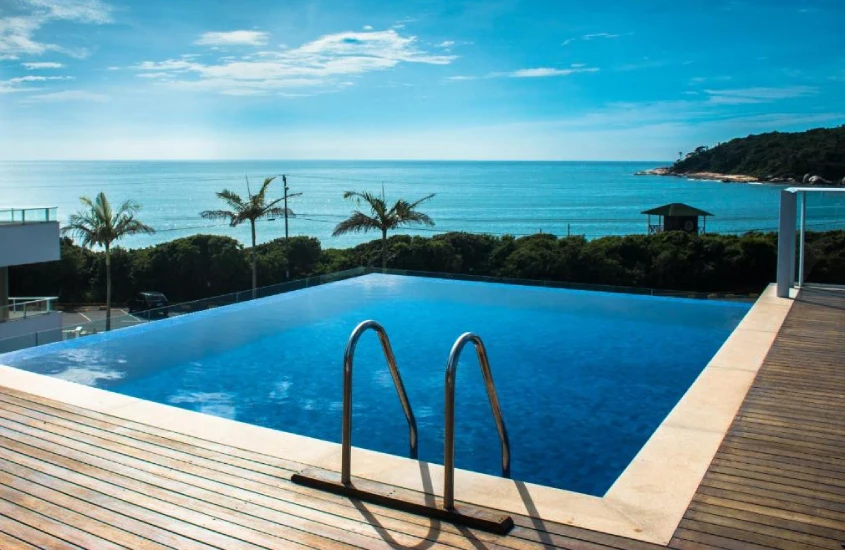 Em um dia ensolarado, piscina infinita com deck de madeira, árvores ao redor e mar na frente