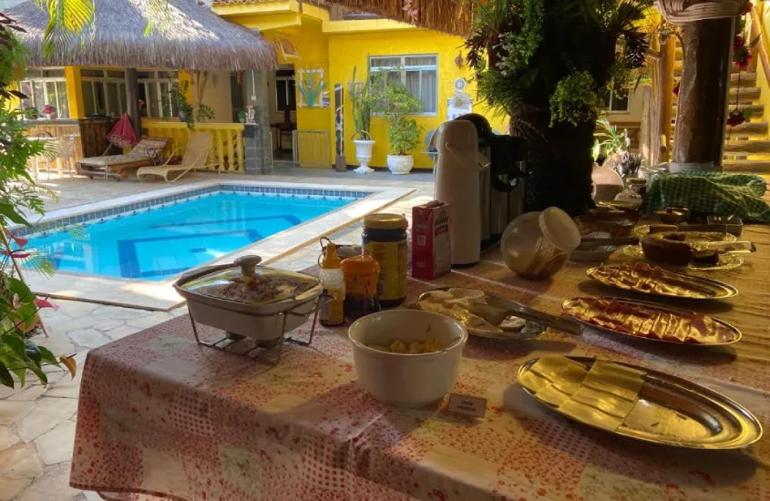 Área de lazer de hotel com mesa de café da manhã, árvores, espreguiçadeiras, guarda-sóis e piscina do lado