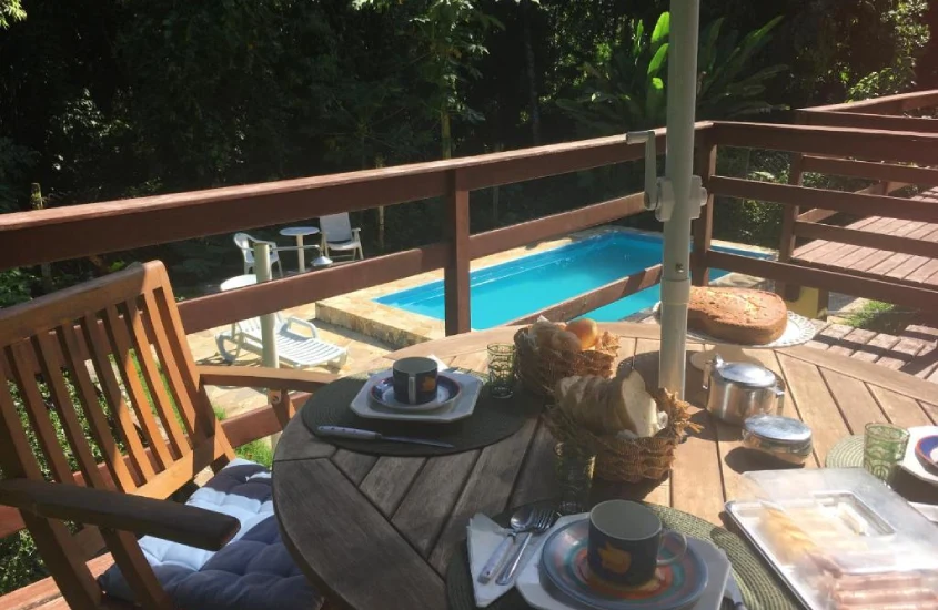 Em um dia de sol, área de lazer de hotel com mesa de café posta, piscina, cadeiras, espreguiçadeiras e árvores ao redor