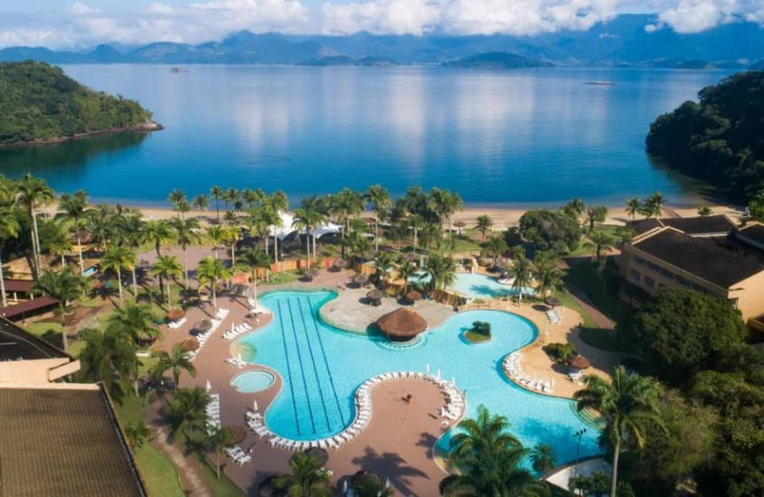 Visão aérea de hotel com piscina, espreguiçadieras, árvores e mar na frente