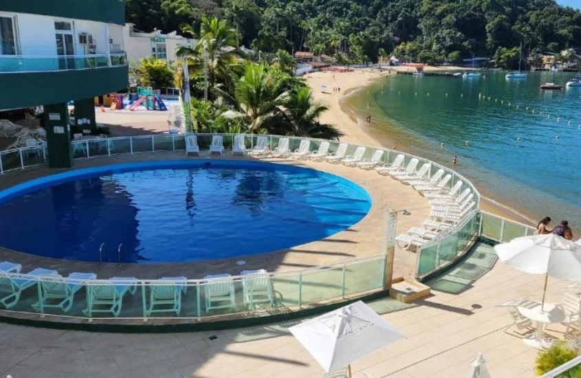 área de lazer de hotel com piscina, espreguiçadeiras, mesas, cadeiras, guarda-sóis, mar, parvores e praia do lado