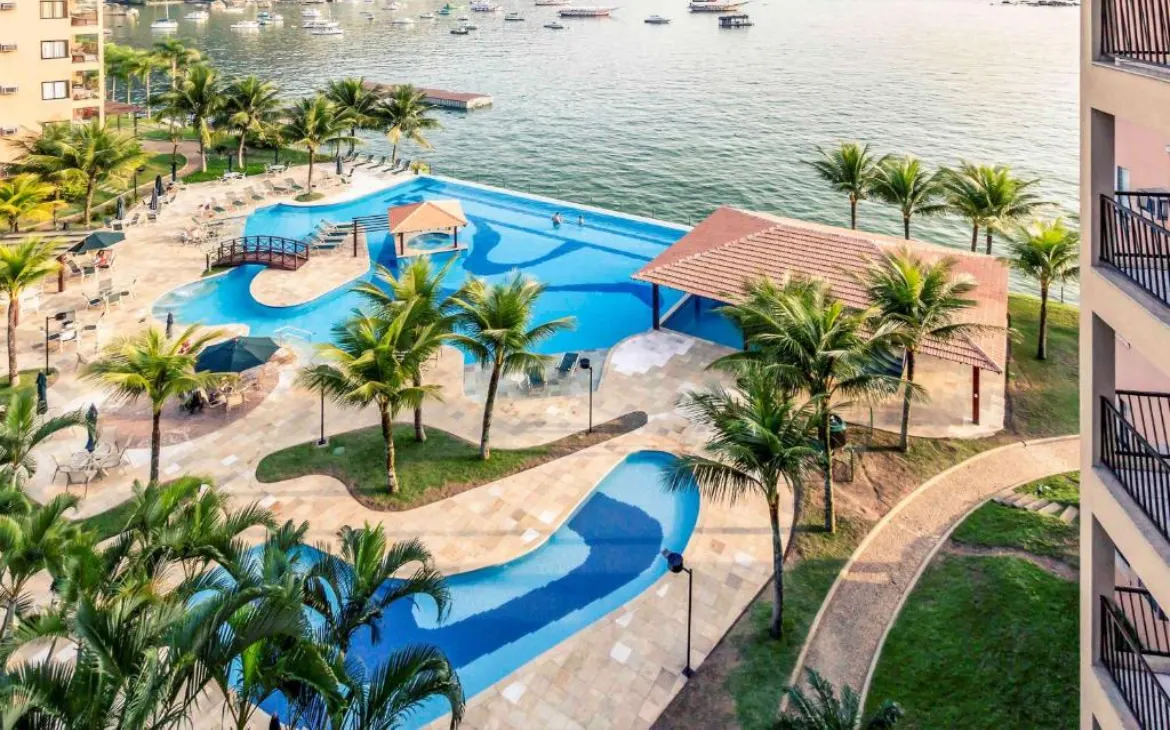 Vista aérea de hotel com piscinas, árvores ao redor e mar do lado