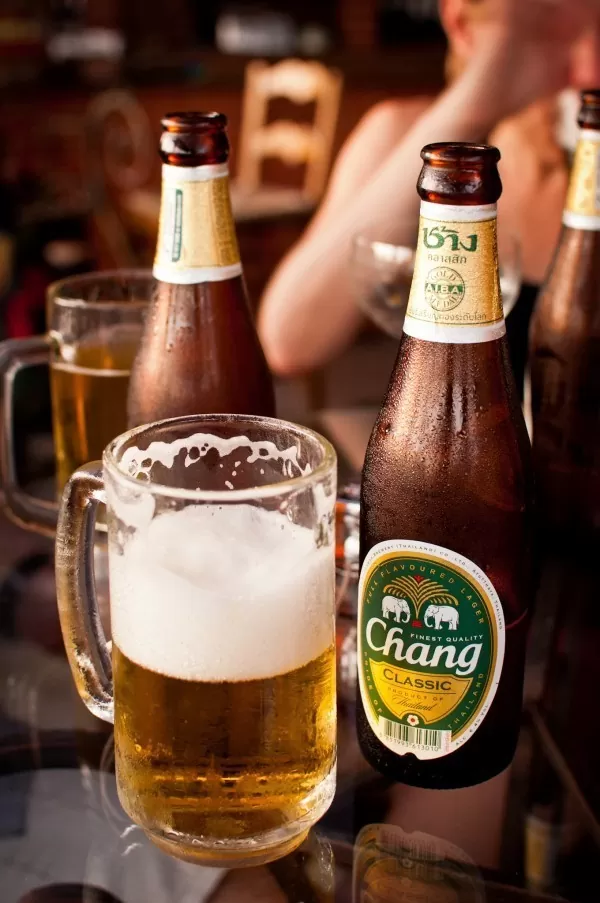 Caneca de vidro com cerveja ao lado de garrafa de vidro marrom de Chang, a cerveja mais vendida da Tailândia