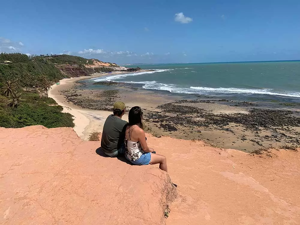 Casal sentado em falésias, durante o dia, obversa mar em Praia da Pipa, um dos lugares para viajar barato