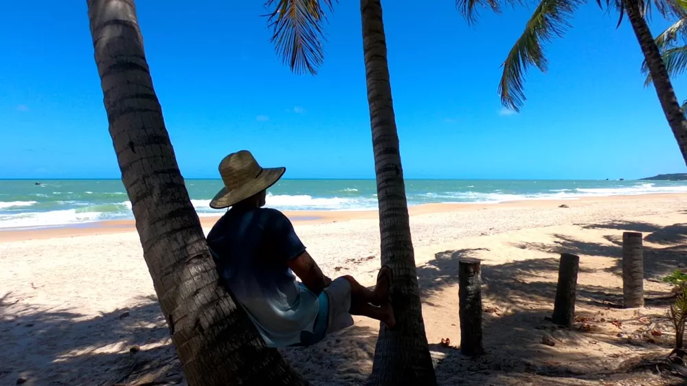 viajante sentado na areia com as pernas esticadas, apoiadas em uma árvore, em praia deserta no litoral Sul, durante o dia