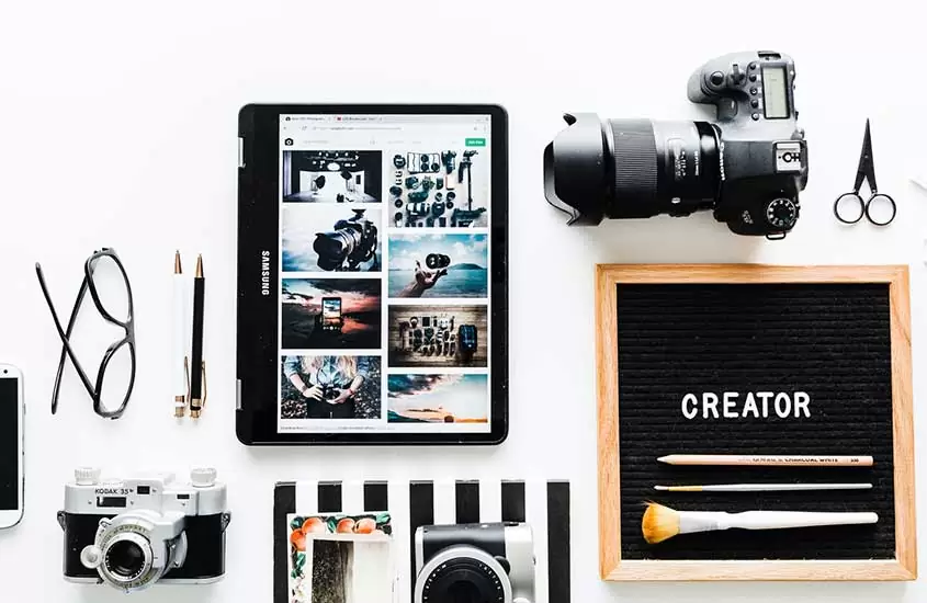 Câmeras fotográficas e tablet em cima de mesa, objetos que podem ser usados por quem quer ganhar dinheiro viajando