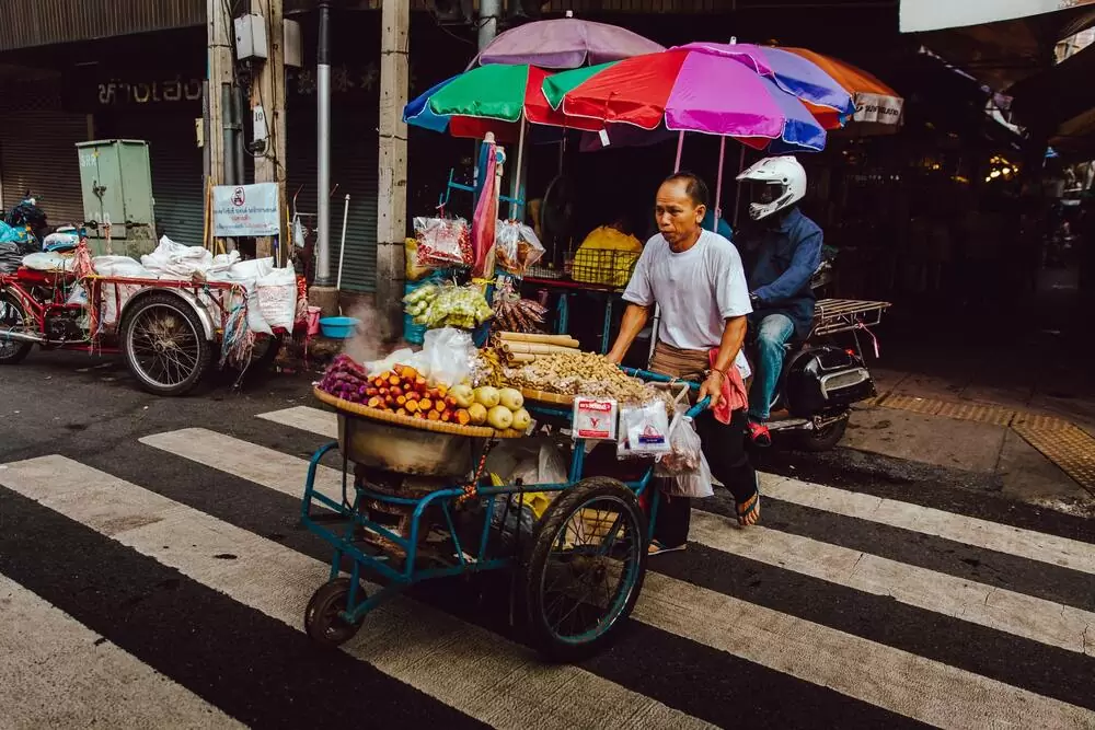 Homem, vendedor ambulante, atravessa rua em Bangkok empurrando carrinho com frutas, durante o dia