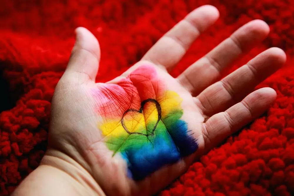 Pessoa com mão estendida onde há desenhado um coração em cima das cores vermelho, amarelo, verde e azul, representando a bandeira LGBT