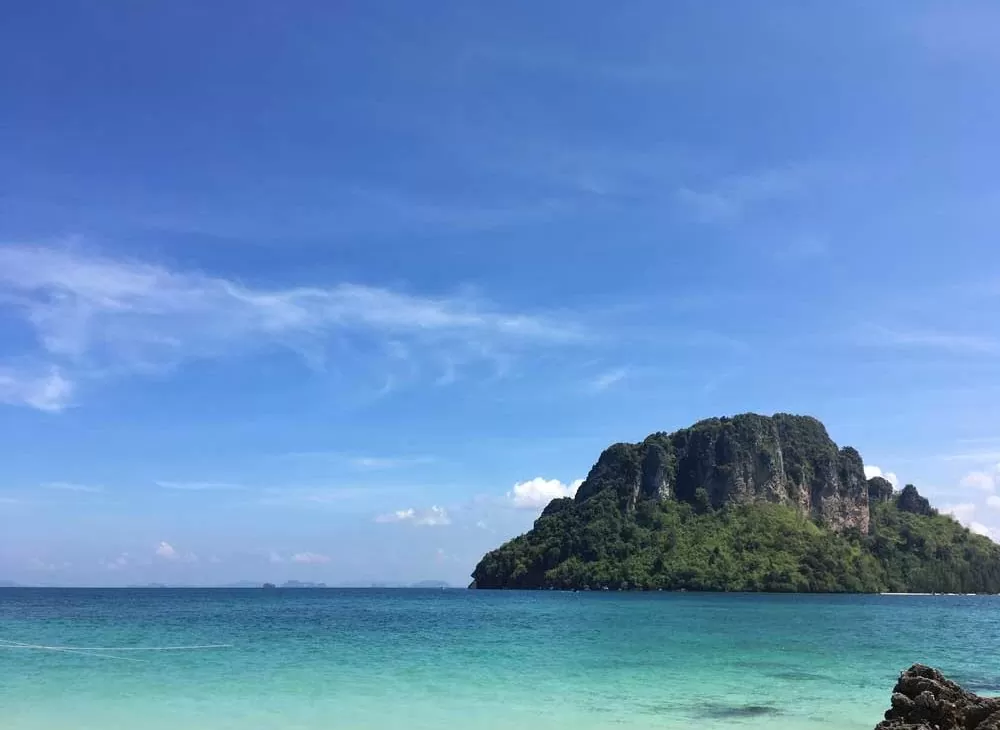 Mar azul, durante o dia, onde ao fundo há uma montanha, em Ao Nang, cidade da Tailândia, um dos lugares para viajar barato.