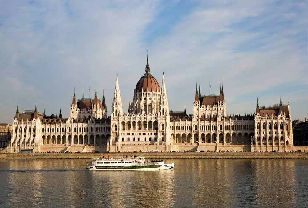 Vista panorâmica de edifícios legislativos de 1885 que fazem parte do Parlamento da Hungria, às margens do rio, onde há um barco, em Budapeste.