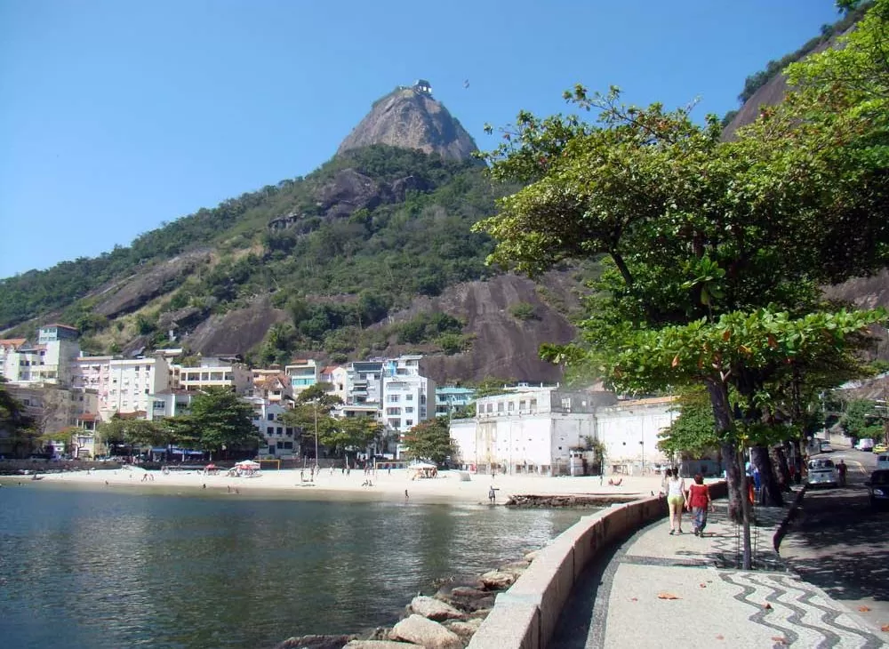 Pessoas caminham em calçada ao lado de muro baixo ao lado da Baía de Guanabara, conhecido como mureta da urca, uma opção para quem busca passeios do rj