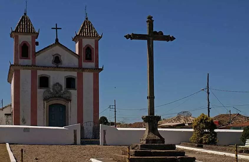 grande cruz em frente a igreja branca com detalhes rosas e porta azul, durante o dia em centro histórico de lavras novas, um dos passeios em minas gerais