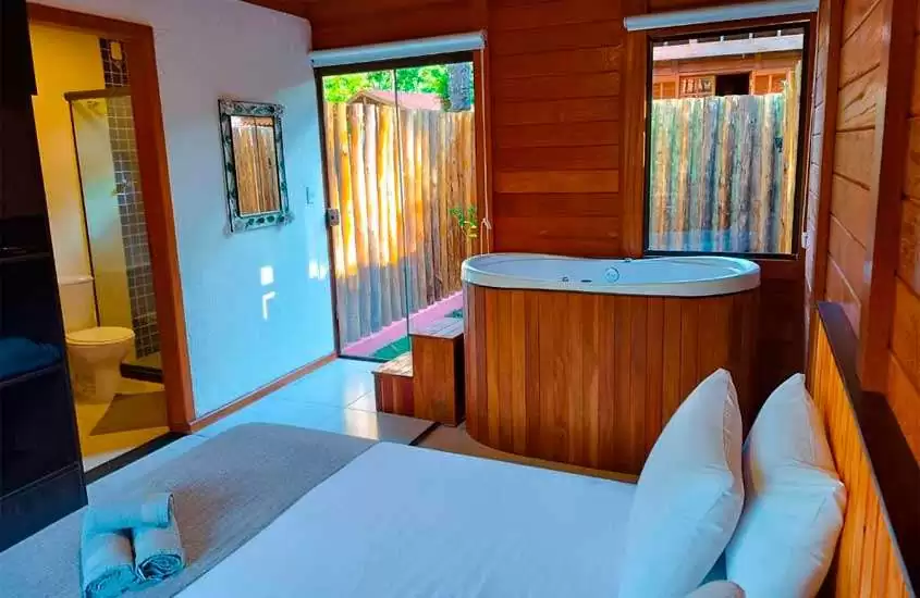 Interior de quarto de hotel com cama de casal, toalhas, banheira de hidromassagem, deck de madeira, janelas grandes, banheiro e TV na parede