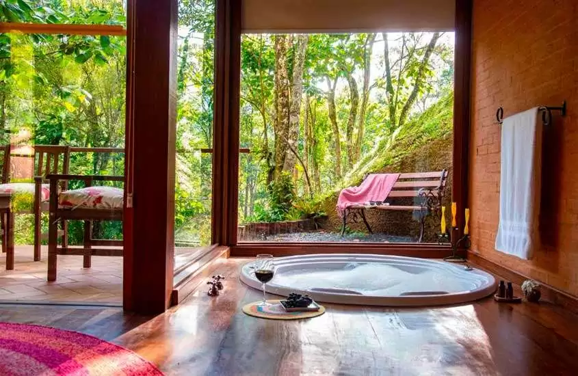 Área de banheira com deck de madeira, toalhas, tapete, varanda com banco e cadeiras de madeira e natureza ao redor