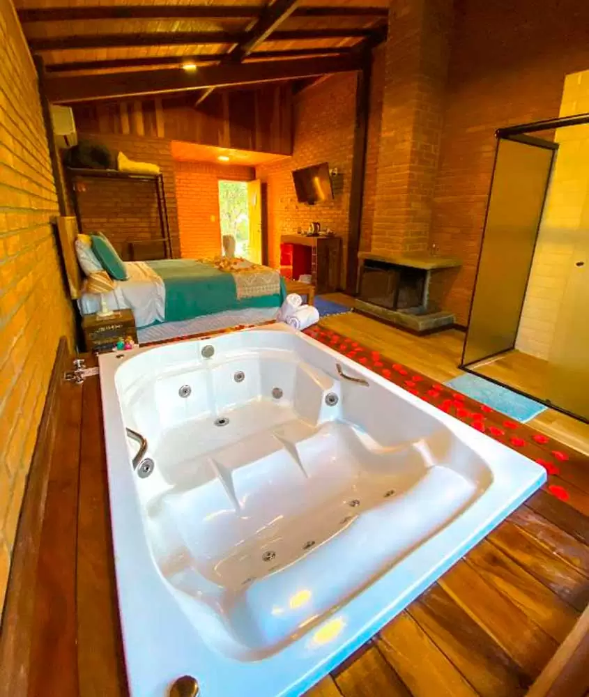 Interior de quarto com cama de casal, banheira de hidromassagem, deck de madeira, lareira, TV, ar condicionado e box