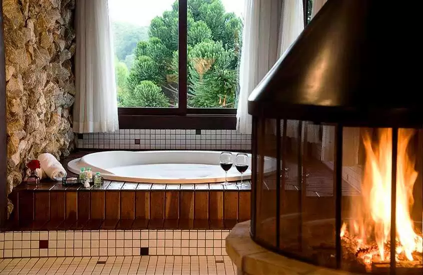 Área de banheira de um hotel onde ficar em visconde de mauá com lareira do lado, parede de pedra, deck de madeira, taças de vinho, toalhas, flores decorativas e janela com vista da natureza