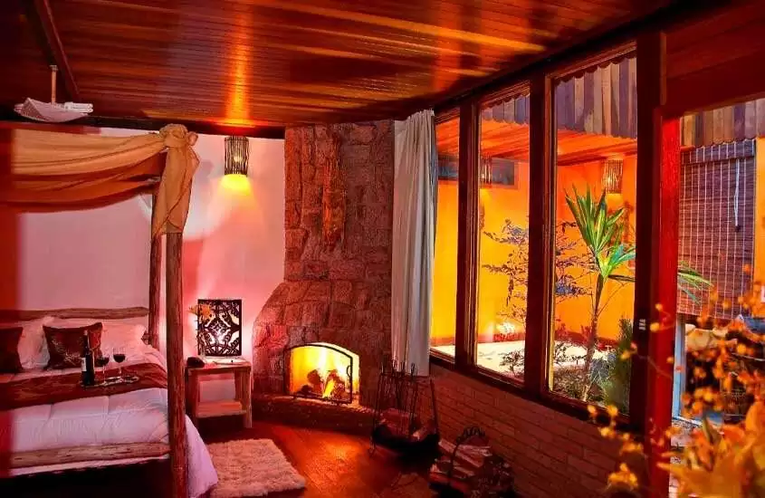 Quarto de um hotel com cama de casal, vinho, tapete, lareira de pedra e janela acortinada com paisagem da varanda