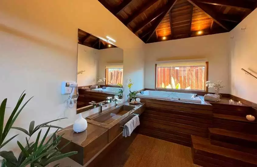 Área de banheira de um hotel onde se hospedar em visconde de mauá com deck de madeira, pia, flores descorativas, espelho, toalhas e janela grande acortinada