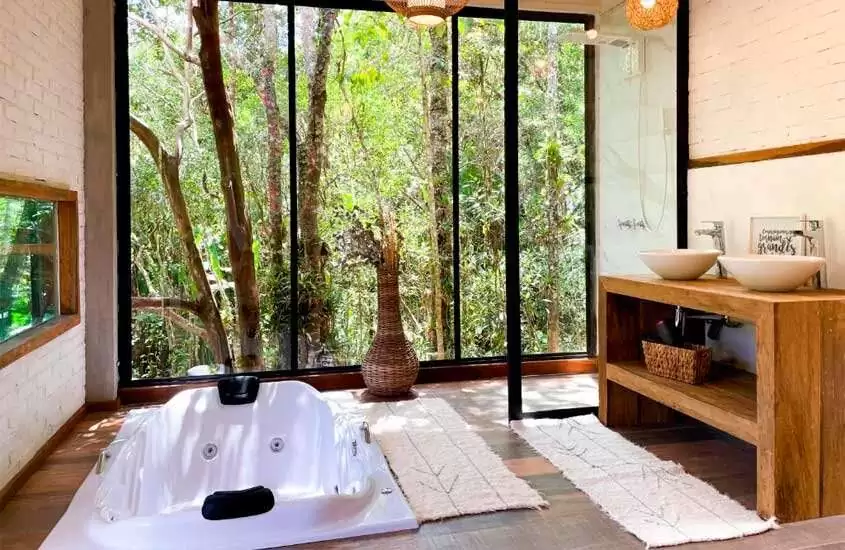 Área de banheira de hotel com banheira de hidromassagem, tapetes, jarro decorativo, pias, chuveiro e janelas grandes com paisagem da natureza