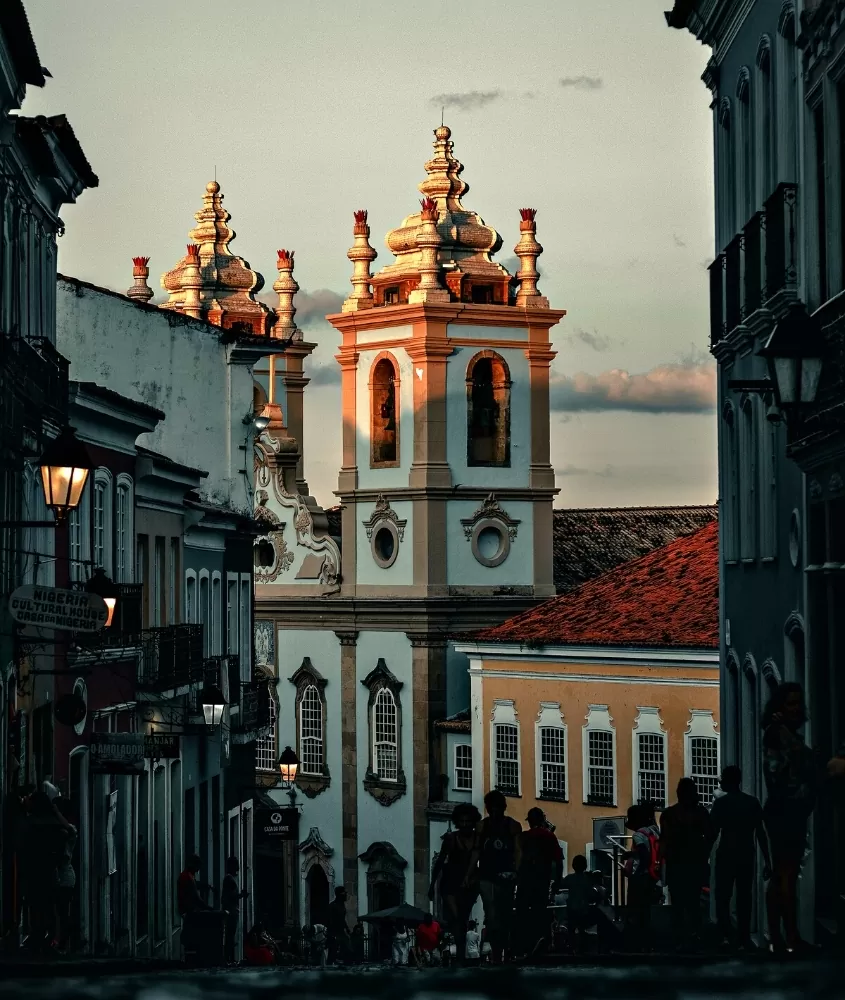 Pessoas caminhando na rua de Pelourinho, em Salvador. Fachada de igreja ao final da rua.