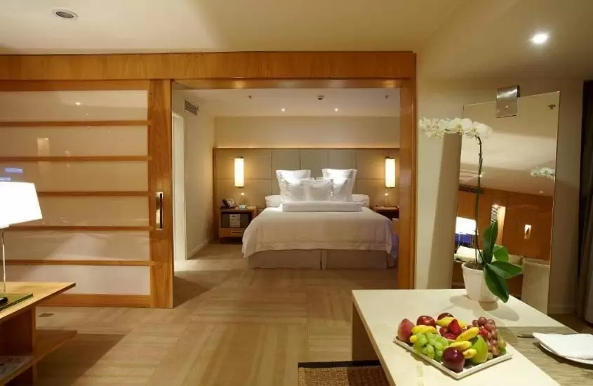 frutas e vaso de flor em cima de mesa e cama de casal ao fundo em quarto de hotel emiliano em são paulo