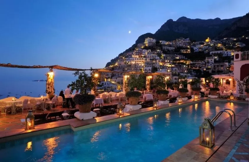 piscina, mesas e cadeiras em área de lazer de le sirenuse, hotel em frente a montanha com casas iluminadas, durante o anoitecer em positano, na itália