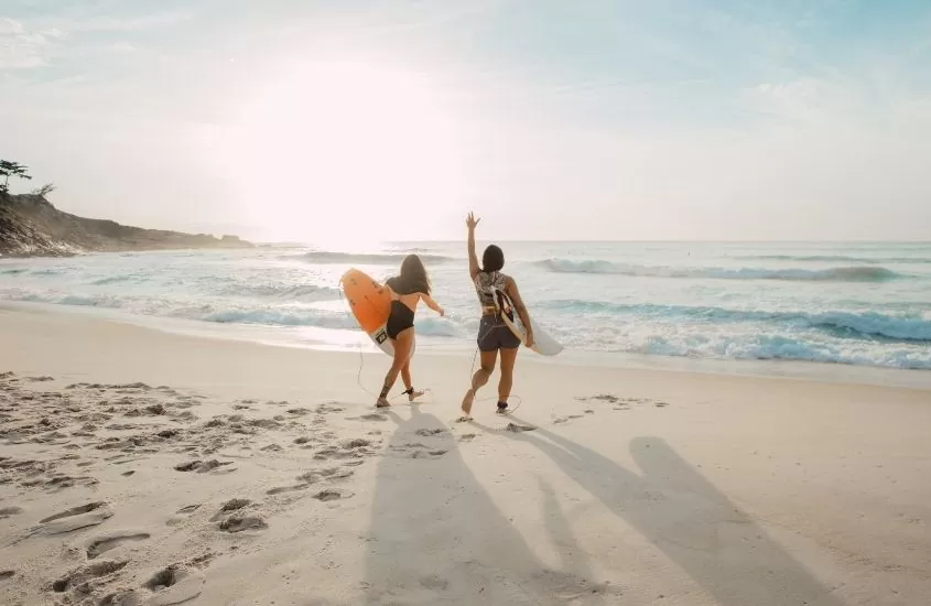 duas mulheres caminham para o mar segurando pranchas de surf
