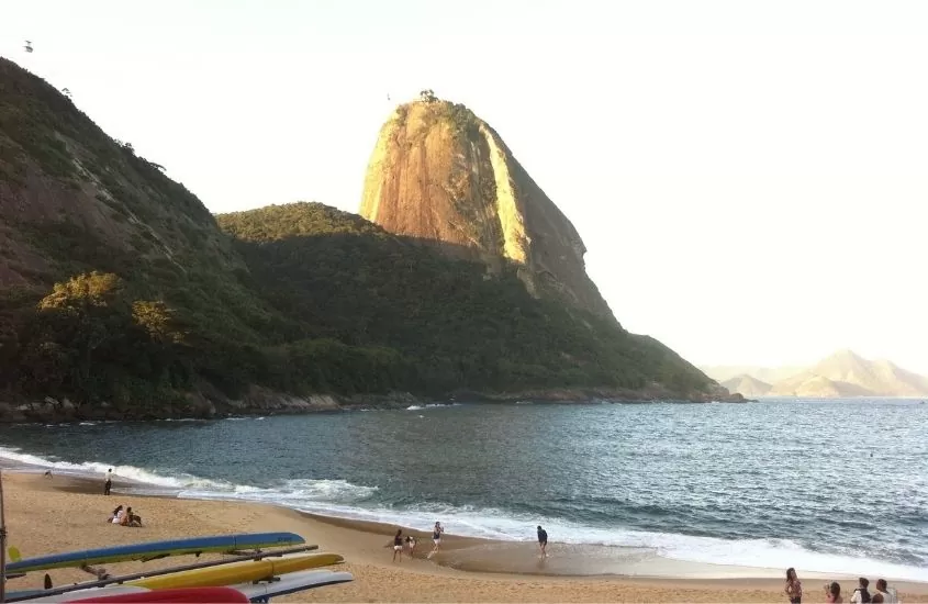 pessoas em areia de Praia Vermelha, onde há a fundo o Morro da Urca, um dos lugares mais bonitos do Rio de Janeiro