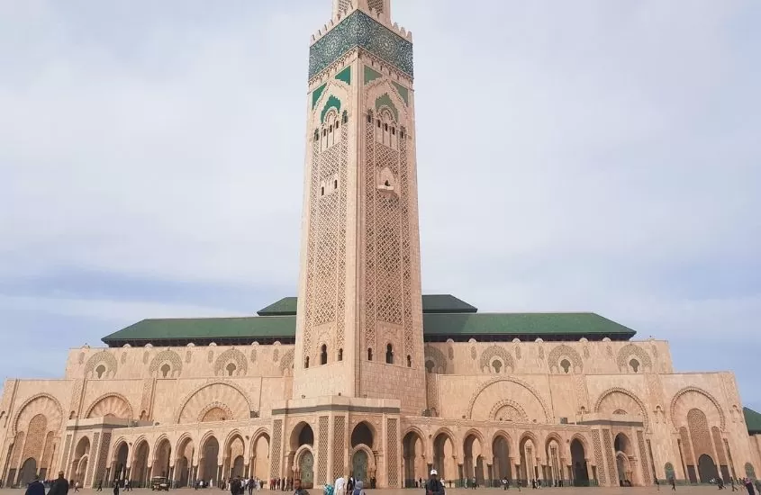 pessoas em frente a construção com 180 metros de altura onde funciona a hassan ii, a maior mesquita do marrocos