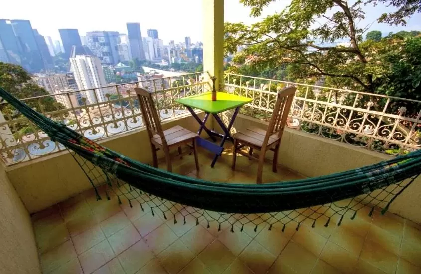 cadeiras, mesa e rede de descanso em varanda com vista para os prédios, durante o dia, em rio forest hostel