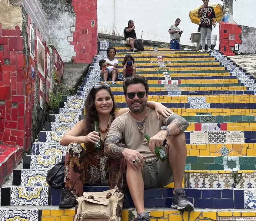 bárbara rocha e vagner alcantelado, casal viajando o mundo, sorriem para foto sentados em escadaria selarón, um dos pontos turísticos de santa teresa rj