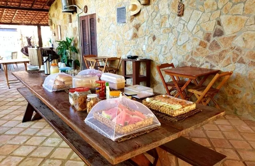 frutas, pães, bolos e biscoitos em cima de mesa de madeira em refeitório de pousada rico atins, uma opção para quem busca pousadas nos lençóis maranhenses