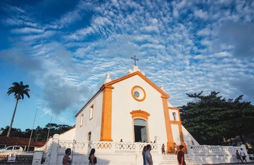 pessoas passam em frente a igreja branca com detalhes amarelo, durante o dia, em Centro de Florianópolis, uma ótima área onde ficar em Florianópolis