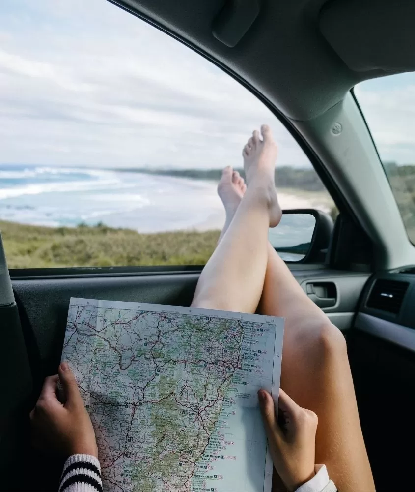 pessoa deitada com as pernas apoiadas em janela de carro, de frente para o mar, segura mapa de papel